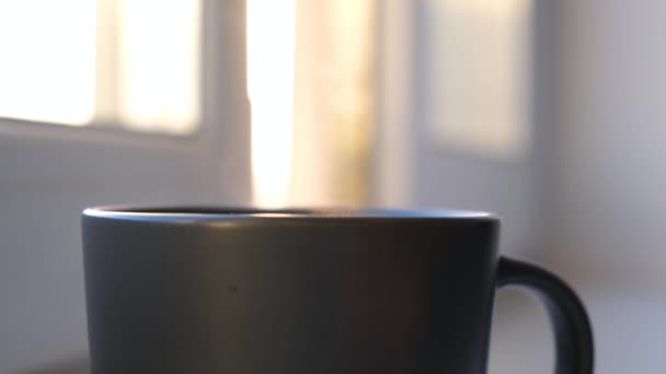 Caneca preta com vapor no fundo da janela ensolarada. Conceito. Close-up de vapor vindo de caneca preta com bebida quente. Conforto em casa com caneca de bebida quente — Vídeo de Stock