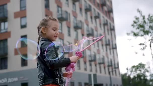 Das süße kleine Mädchen spielt draußen auf dem Spielplatz mit riesigen Seifenblasen, eine glückliche Kindheit. Handeln. Kaukasisches Mädchen macht riesige Seifenblasen, die hoch in der Luft schweben. — Stockvideo