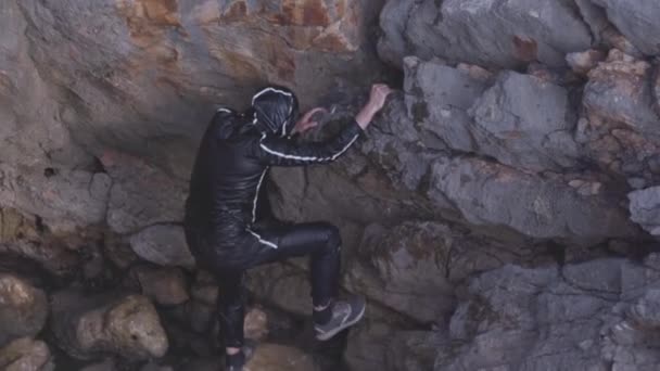 山に登るしようとしている彼の服を完全に濡れている静かな海岸に男。株だ。黒のウェットスーツの男が海によって大きな崖に到達しようとしている. — ストック動画