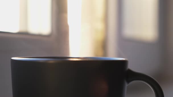 Een kopje warme koffie of thee en de stoom die opstijgt in de lucht. Concept. close-up van zwarte beker met warme drank staan in de voorkant van het raam thuis op wazig achtergrond. — Stockvideo