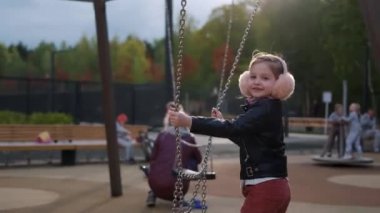 Güzel küçük kız salıncakta sallanıyor, yüzünde mutlu bir gülümsemeyle. Başla. Siyah ceketli, pembe kulaklıklı, dışarıda eğlenen bir kızın yan görüntüsü..