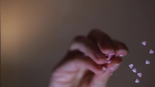Kobiece dłonie rzucające różowe konfetti w kształcie serca na płaską przezroczystą powierzchnię. Koncepcja. Widok z dołu żeńskiej dłoni kładącej małe serduszka na szklanym stole. — Wideo stockowe