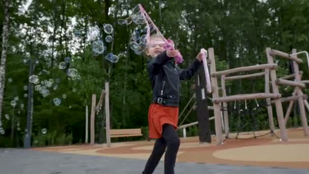 Bonne fille souriante jouant avec des bulles de savon géantes. L'action. Enfant femelle soufflant de grandes bulles, courant et s'amusant à l'extérieur à l'aire de jeux moderne de la ville. — Video