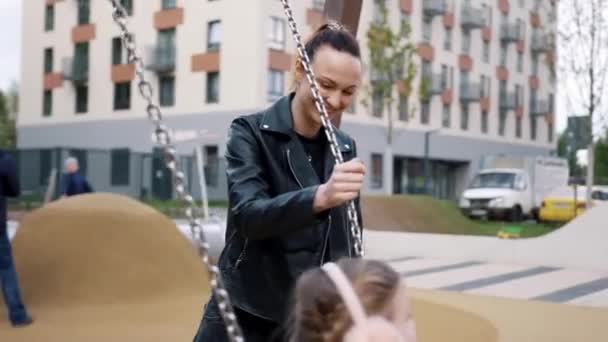 Hermosa madre joven balanceando a su niña en columpios en el patio de recreo con una sonrisa feliz en sus rostros. Acción. Niña vistiendo chaqueta negra divirtiéndose al aire libre en columpios y una pirámide de cuerda . — Vídeos de Stock