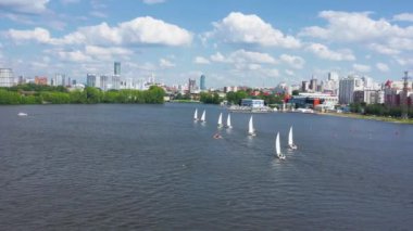 Şehrin arka planında yüzen yatların en üst görüntüsü. Video kaydı. Yelkenli yatları olan bir göl manzarası ve şehir ve gökyüzü olan bir ufuk. Şehrin çarpıcı manzaralı yelkenli tekneleri yarışı.