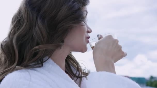 Aantrekkelijke vrouw in witte jas drinkt koffie staande op balkon. Een video. Zelfvoorzienende vrouw rust in dure hotel koffie drinken in de ochtend op het balkon — Stockvideo