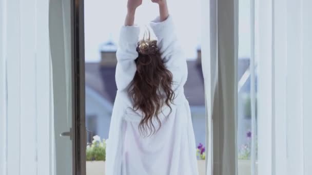 Aantrekkelijke vrouw strek op balkon. Een video. Achteraanzicht van een mooie vrouw in witte jas die zich uitstrekt op een balkon op zonnige ochtend. Aantrekkelijke vrouw op vakantie in het buitenland — Stockvideo