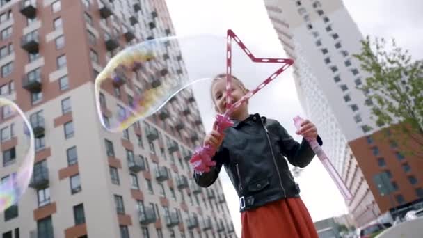 Retrato de un niño creando burbujas de jabón cerca del edificio de varios pisos. Acción. La linda niña está jugando con enormes burbujas de jabón al aire libre, concepto de infancia feliz . — Vídeo de stock