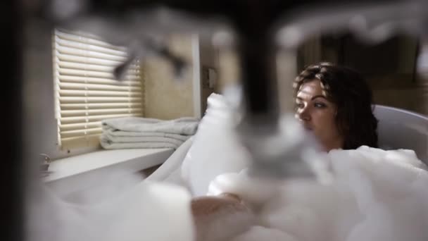 Сексуальна жінка лежить у бульбашковій ванні і дивиться у вікно. Відео. Красива жінка відпочиває в розкішній бульбашковій ванні біля вікна. Розслабляється в гарячій ванні з піною — стокове відео