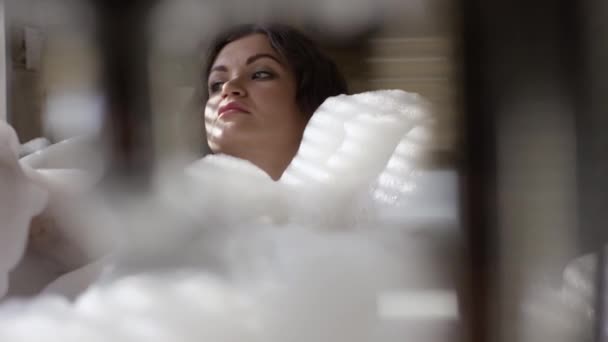 Сексуальная женщина лежит в пенной ванне и смотрит в окно. Видео. Красивая женщина расслабляется в роскошной ванне с пеной за окном. Отдых в гидромассажной ванне с пеной — стоковое видео