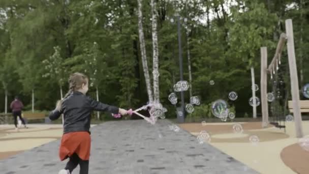Chica rubia activa con una trenza que crea burbujas mientras corre por el parque de la ciudad. Acción. Adorable niña emocionada jugando con burbujas de jabón en el patio de recreo, vista trasera . — Vídeos de Stock