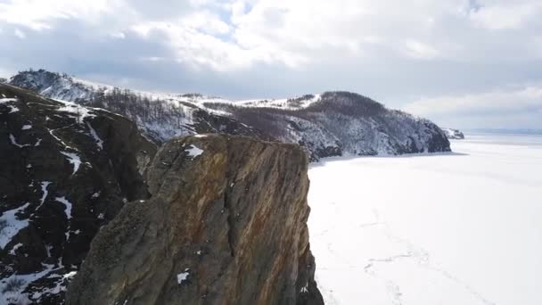 Uma rocha íngreme e a margem de um lago congelado. Clipe. Vista aérea do penhasco íngreme e montanhas cobertas por árvores carecas e o lago sob o gelo e neve, paisagem natureza inverno . — Vídeo de Stock