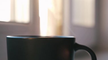 Pencere pervazında kahve ya da çay fincanı. Kavram. Karanlık bardaktaki sıcak içeceği kapat. Buhar yükseliyor ve arka planda güneş ışığı parlıyor..