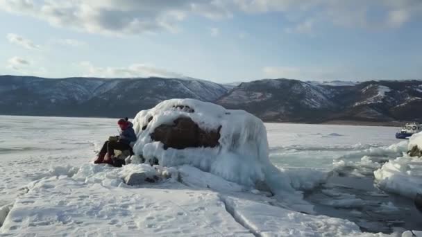 Weibchen auf der eisigen und schneebedeckten Oberfläche des Sees unterwegs. Clip. Luftaufnahme einer Frau, die im Winter auf einem Stein am zugefrorenen Baikalsee sitzt und eine stählerne Zungentrommel spielt. — Stockvideo