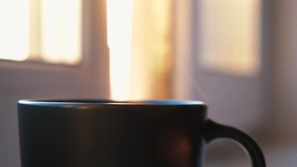Kávé vagy teáscsésze az ablakpárkányon. Fogantatás. Közel a forró ital a sötét bögrében a gőz emelkedik, és a napfény ragyog az ablakon keresztül a háttérben. — Stock videók