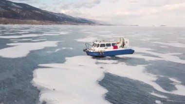 Hovercraft düz kar ve buz yüzeyinde ilerliyor. Şarjör. Kurtarma aracının hava manzarası kış mevsiminde donmuş gölün dağlık kıyısı boyunca ilerliyor..