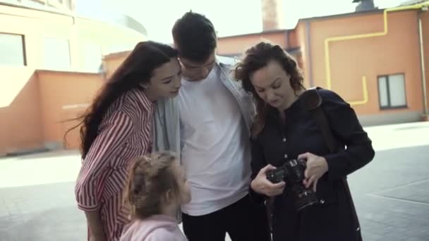 Kobieta i para z uroczą dziewczynką oglądającą zdjęcia przed kamerą. Akcja. Fotograf pokazujący zdjęcia szczęśliwej młodej rodzinie na świeżym powietrzu w letni dzień. — Wideo stockowe