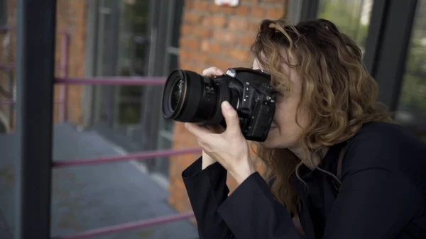 Retrato de fotógrafa com câmera profissional. Acção. Vista lateral da mulher caucasiana com cabelo encaracolado tirar fotos em sua câmera no fundo da parede de tijolo vermelho . — Fotografia de Stock