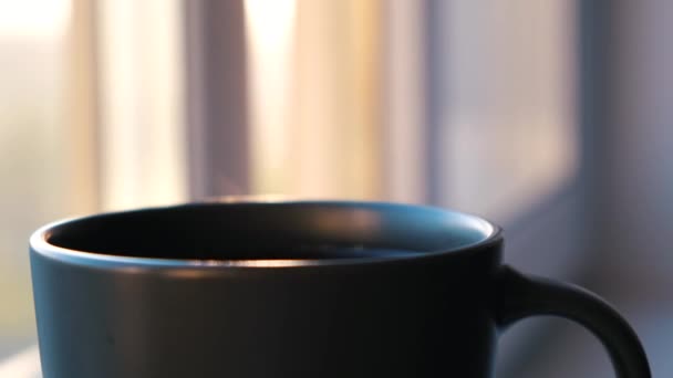 Una taza de bebida caliente cerca de la ventana en un día soleado. Concepto. Primer plano de la taza con la bebida caliente con el aumento de vapor sobre fondo borroso, atmósfera de la mañana . — Vídeos de Stock