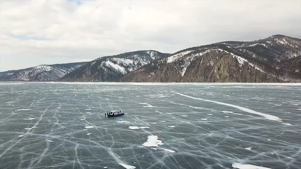 La gente viaja en el hielo del lago congelado Baikal cerca de un aerodeslizador seguro y cómodo Khivus. Clip. Vista aérea de un grupo de turistas caminando sobre el hielo grueso de lago increíble, concepto de turismo extremo . — Foto de Stock