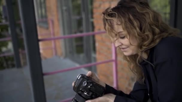 Portret van vrouwelijke fotograaf met professionele camera. Actie. Zijaanzicht van Kaukasische vrouw met krullend haar het nemen van foto 's op haar camera op rode baksteen muur achtergrond. — Stockvideo