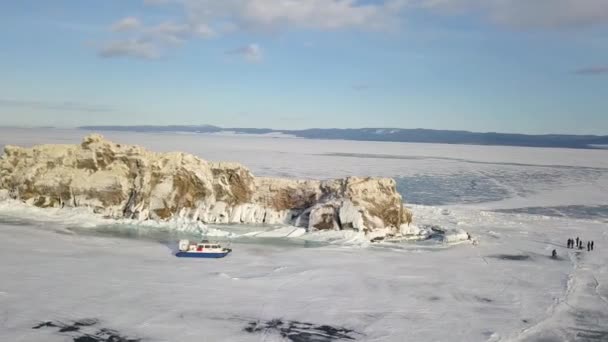 Die Menschen bewegen sich auf dem Eis des zugefrorenen Baikalsees in der Nähe sicherer und komfortabler Hivus-Luftkissenboote. Clip. Luftaufnahme einer Gruppe von Touristen zu Fuß auf dickem Eis des erstaunlichen Sees, Extremtourismus-Konzept.