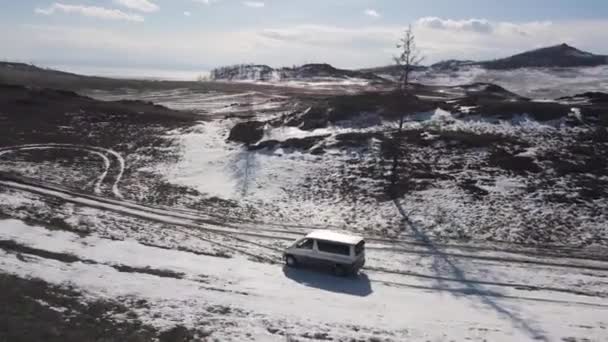 Aerial view of the vehicle driving in winter high hills on cloudy sky background. Clip. Hiking in cold weather, minivan driving in mountains covered by trees, yellow grass, and snow. — Stock Video