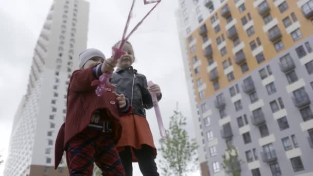 Schöne Mädchen blasen Blasen, Ansicht von unten. Handeln. Weibliche kaukasische Kinder in warmen Kleidern, die im Frühling auf mehrstöckigen Gebäuden viele Seifenblasen erzeugen. — Stockvideo