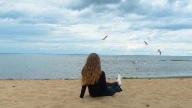 Gölün kumlu kıyısında oturan kadının arka görüntüsü. Kavram. Kumda oturan güzel genç kadın bulutlu gökyüzü ile deniz ufkunun tadını çıkarıyor.