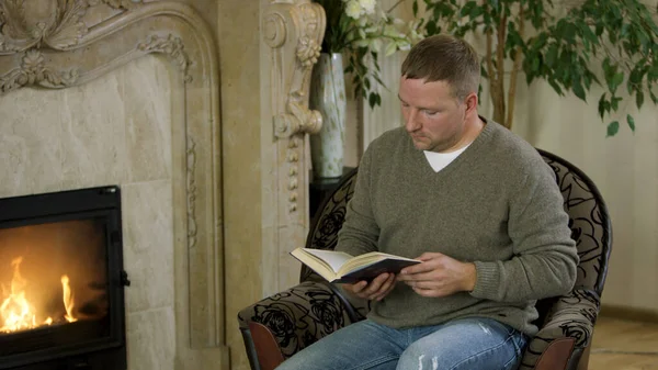 Man sitting in a chair,reading a book in front of fireplace. Video. Butterfy flying near the male wearing pullover and jeans sitting at home by the fire and reading.