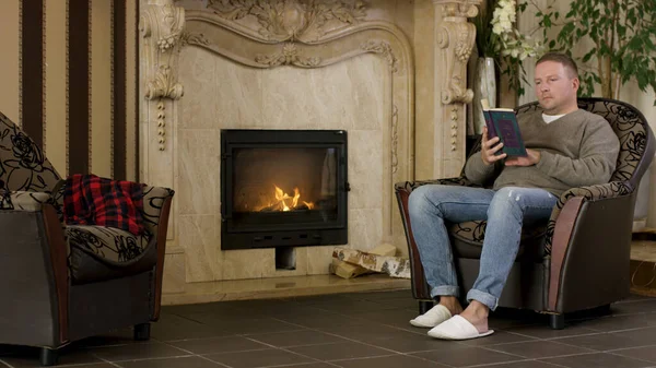 Young man sitting in a chair at home, reading a book in front of fireplace. Video. Male wearing pullover and jeans sitting at home by the fire and reading.