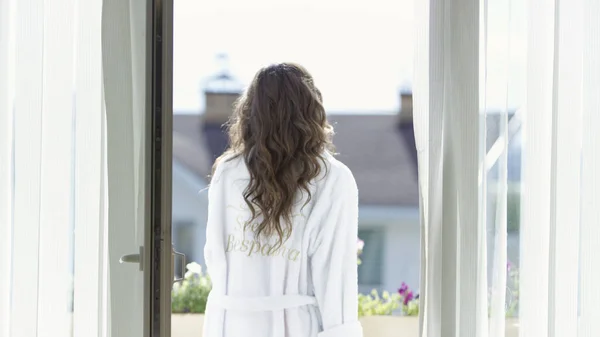 Egy fiatal nő vakáción sétál az erkélyen fehér fürdőköpenyben. Videó. Hátsó kilátás egy fiatal barna nő élvezi nyári napsütés az erkélyen egy frottír köntös. — Stock Fotó