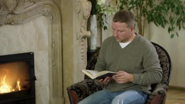 Sandalyede oturan, şöminenin önünde kitap okuyan bir adam. Video kaydı. Erkeğin yanında uçan, kazaklı, kot pantolonlu, şöminenin yanında oturan ve kitap okuyan..