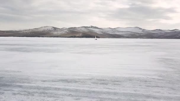 Ohromující přírodní krajina sibiřské divoké přírody. Klip. Letecký pohled na ledový člun na ledě a sněhu jezera Bajkal v pozadí ostrova Olkhon v zimním oblačném dni. — Stock video