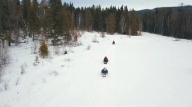 Kar araçlarının hava görüntüsü ormanın yakınındaki karlı bir patikada ilerliyor. Şarjör. Kış ekstrem spor anlayışı, insanlar yoğun gökyüzünün altındaki ağaçlar boyunca kar araçlarına biniyor..