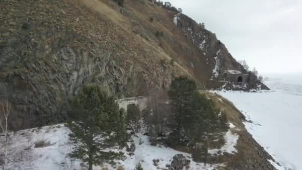 Vue aérienne d'un bâtiment en ruine situé sur le versant d'une montagne près du lac gelé. Clip. Survolant une maison en brique abandonnée construite sur une colline boisée en hiver près d'une rivière glacée. — Video
