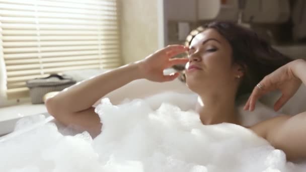 Молодая привлекательная брюнетка расслабляется и принимает ванну с пеной. Видео. Женщина с кудрявыми темными волосами принимает ванну в доме у окна. — стоковое видео