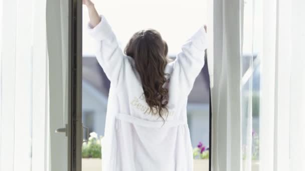 Młoda kobieta na wakacjach spacerująca po balkonie w białym szlafroku. Nagranie. Widok na tył młodej brunetki cieszącej się letnim słońcem na balkonie w szlafroku frotte. — Wideo stockowe