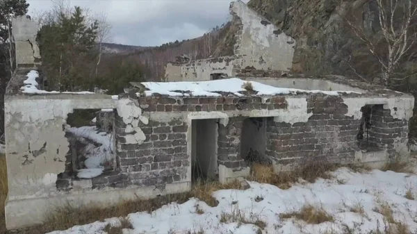 Aerial view of a ruined building located on the slope of a mountain near the frozen lake. Clip. Flying over abandoned brick house built on forested hill in winter season near icy river.