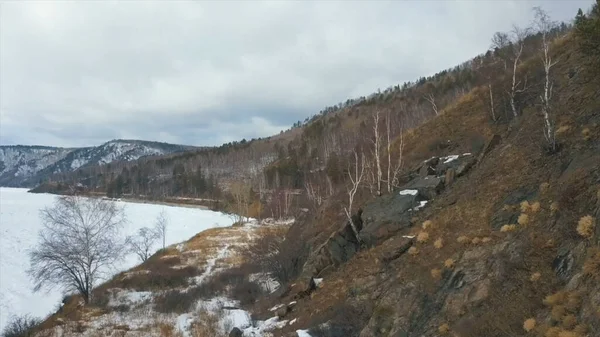 Belle vue sur la rivière enneigée d'hiver et la pente rocheuse avec herbe jaune flétrie. Clip. Aérienne de la surface glacée de la rivière entourée de montagnes enneigées par une journée d'hiver. — Photo