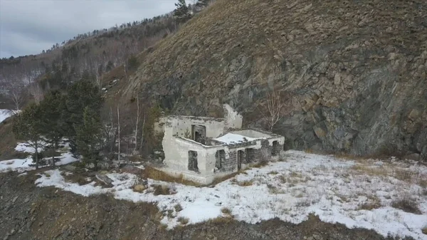 Vista aérea de um edifício arruinado localizado na encosta de uma montanha perto do lago congelado. Clipe. Voando sobre casa de tijolo abandonada construída na colina florestada na temporada de inverno perto do rio gelado . — Fotografia de Stock