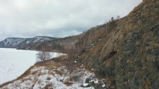 Bella vista sul fiume invernale innevato e sul pendio roccioso con erba gialla appassita. Clip. Aerea di superficie ghiacciata del fiume circondata da montagne innevate in una giornata invernale. — Video Stock