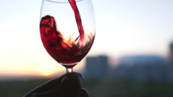 Poner bebida alcohólica en el vaso. Acción. Primer plano de verter vino tinto en el elegante vidrio transparente sobre un fondo borroso de la puesta de sol de verano. — Vídeos de Stock