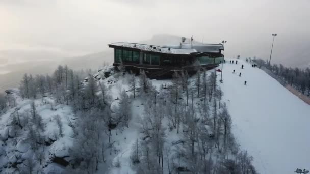 Aerial view of the ski resort with snowy mountain slopes and winter trees. Stock footage. Upper station of cableway and a group of people going skiing and snowboarding down the hill. — Stock Video