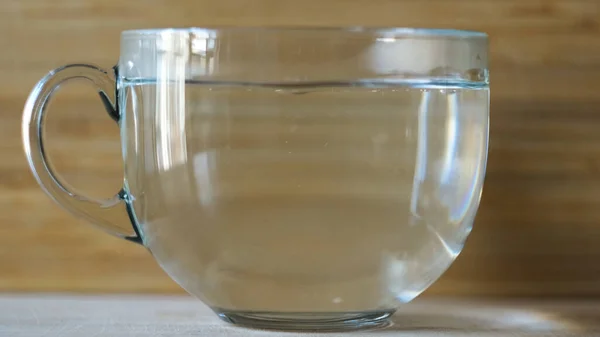 Heißes Wasser in eine große transparente Tasse auf verschwommenem Holzgrund gießen. Konzept. Seitenansicht einer leeren Tasse und der Prozess des Porierens von heißem Wasser für das morgendliche Heißgetränk. — Stockfoto