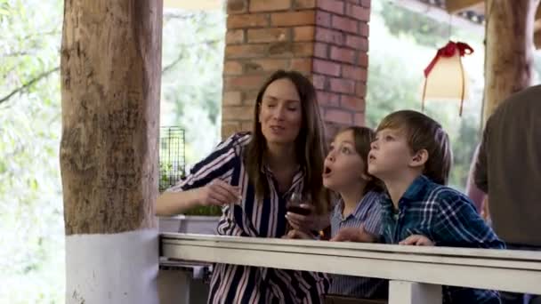 Madre con una copa de vino mostrando algo a sus hijos. Vídeo. Celebración, vacaciones y concepto de fiesta, familia feliz divirtiéndose en la fiesta en una casa de campo . — Vídeos de Stock