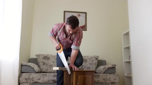 Carpintero cortando una tabla de madera con sierra de mano en casa en la sala de estar. Vídeo. Joven manitas confiado usando sierra durante la reparación de apartamentos . — Vídeo de stock