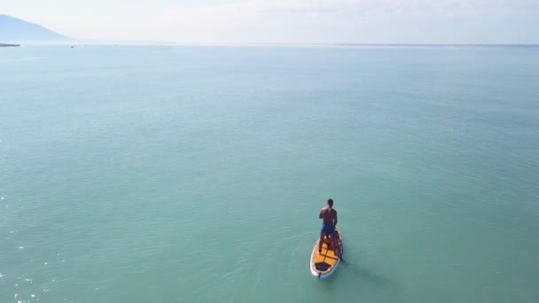 Vista aérea do drone de um homem que pratica stand up paddle ou SUP em mar tropical exótico. Vídeo. Voando sobre o mar azul-turquesa calma e um homem de pé em uma prancha de jantar em montanhas e fundo céu nublado . — Vídeo de Stock