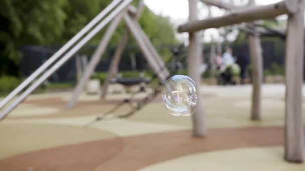 Gros plan de bulles de savon volant lentement sur le fond flou d'une aire de jeux. L'action. de belles bulles de savon à l'extérieur dans la rue d'une ville moderne. — Video