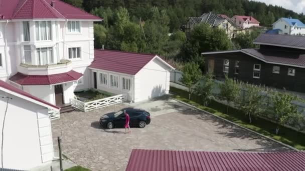 Vue aérienne d'une femme en robe rose élégante rentrant à la maison dans son chalet de campagne. Vidéo. Femme d'affaires verrouillant sa voiture chère et allant vers belle maison blanche de trois étages. — Video